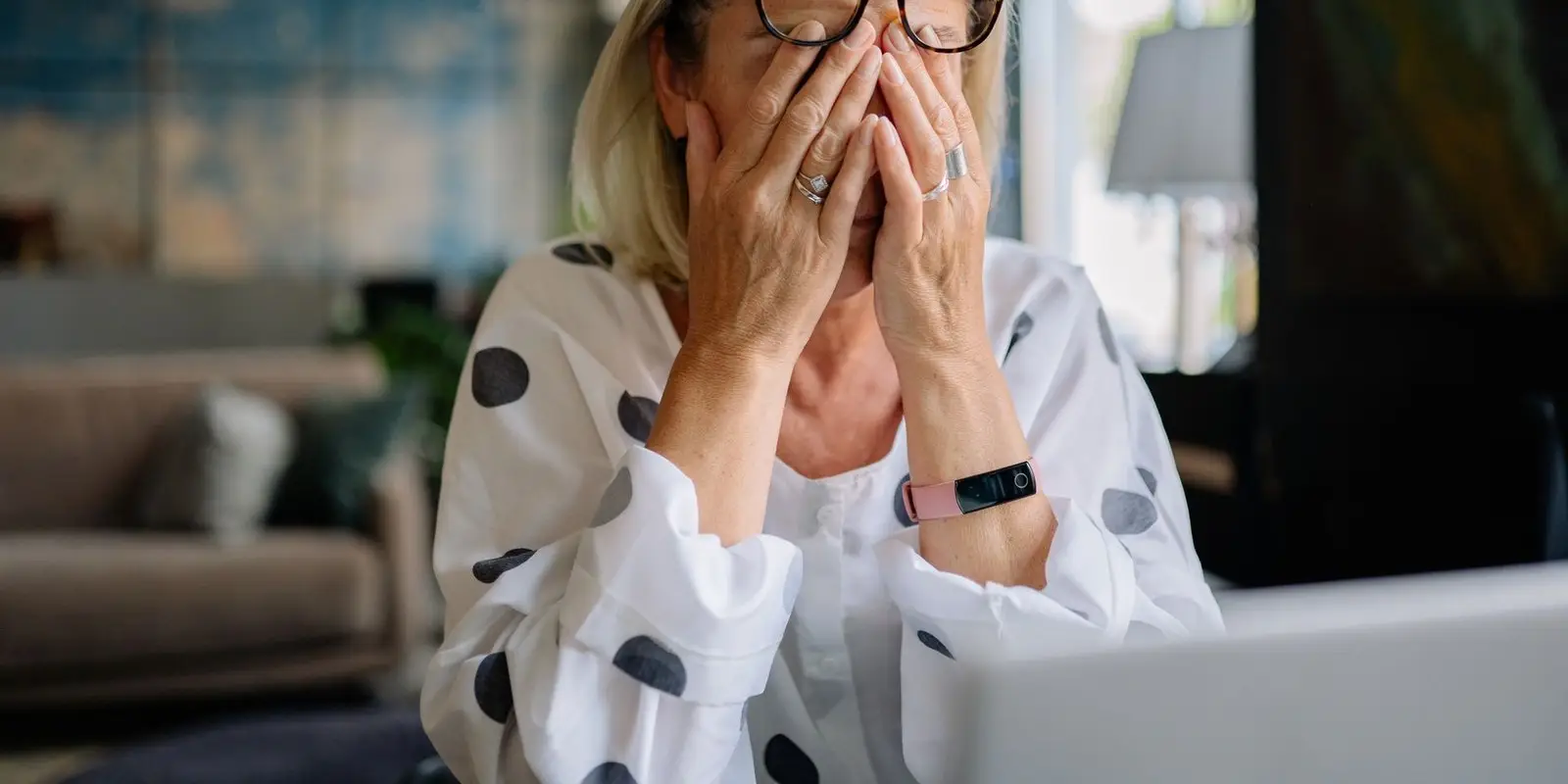 Una donna sui 55-60 anni, seduta alla scrivania di casa, si copre il viso con le mani in un momento di stanchezza.